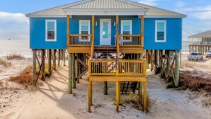 Een blauw strandhuis met een houten terras en een deur, gebouwd op palen boven een zandstrand.