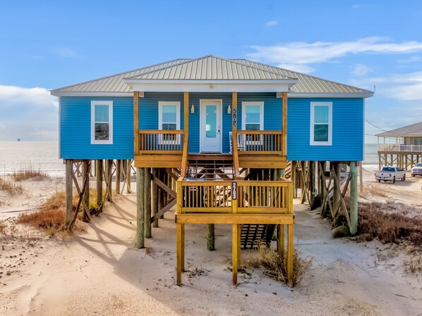 Una casa playa azul con terraza de madera y una puerta, construida sobre pilotes sobre una playa arenosa.
