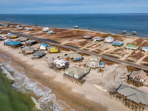 Vista aérea de una zona costera con casas sobre pilotes, una playa y un muelle que se adentra en el agua.