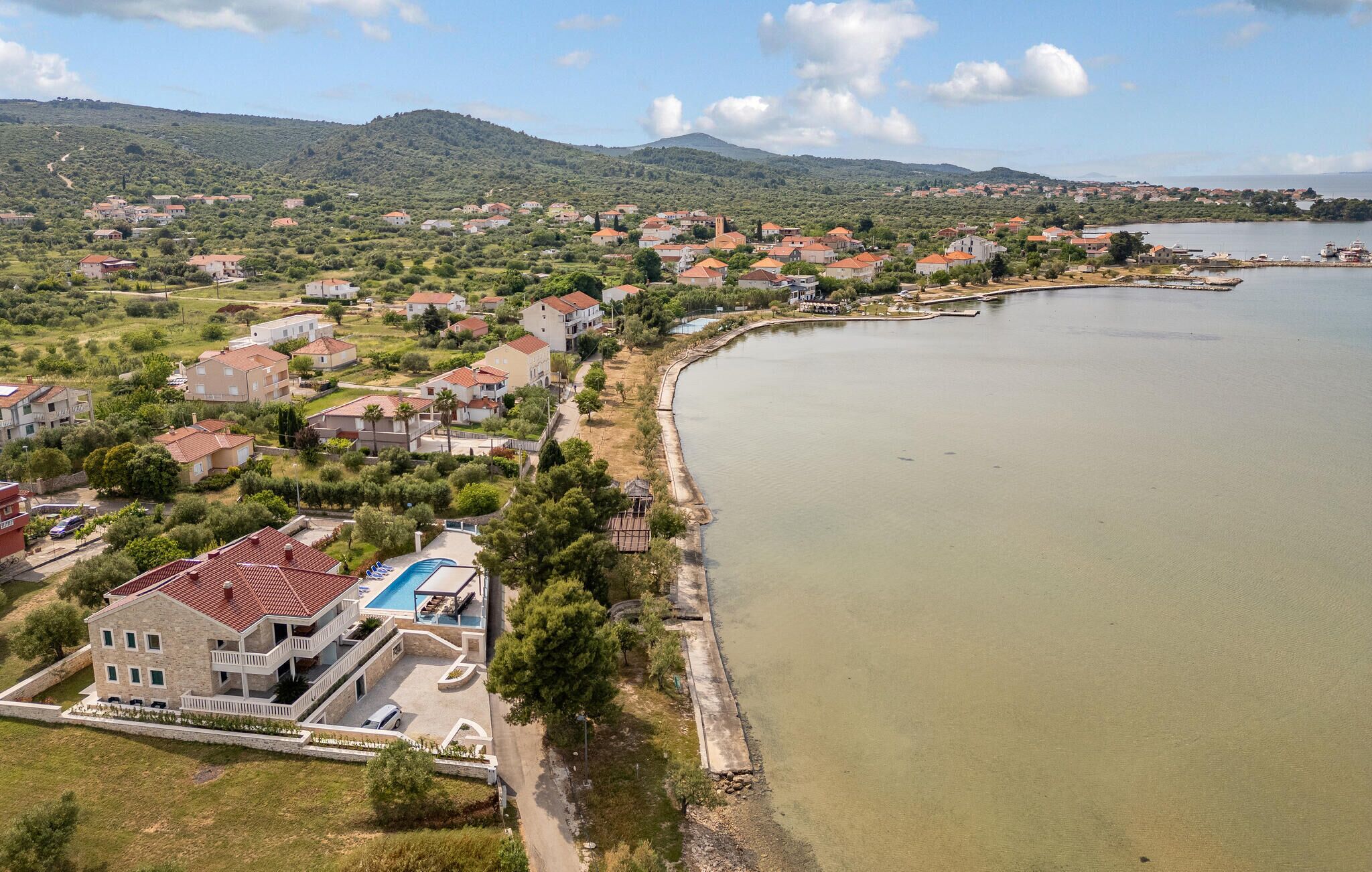 A coastal residential area with houses along a curved shoreline, a swimming pool, and a hilly backdrop.