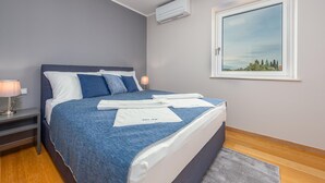 A neatly made bed with a blue and white bedspread, a gray headboard, and a window with a view of trees and sky. - Cozy apartment in Mrljane (Mrljane)