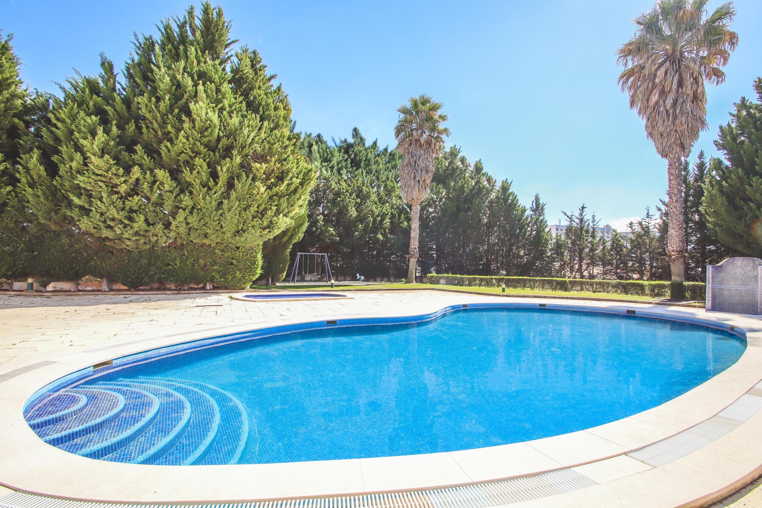 Une piscine dont le bord est revêtu de carrelage, entourée d’arbres et surplombée d’un ciel dégagé.