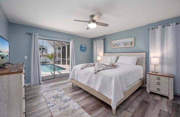 A bedroom with a bed, bedside tables, a ceiling fan, and a view of a swimming pool.