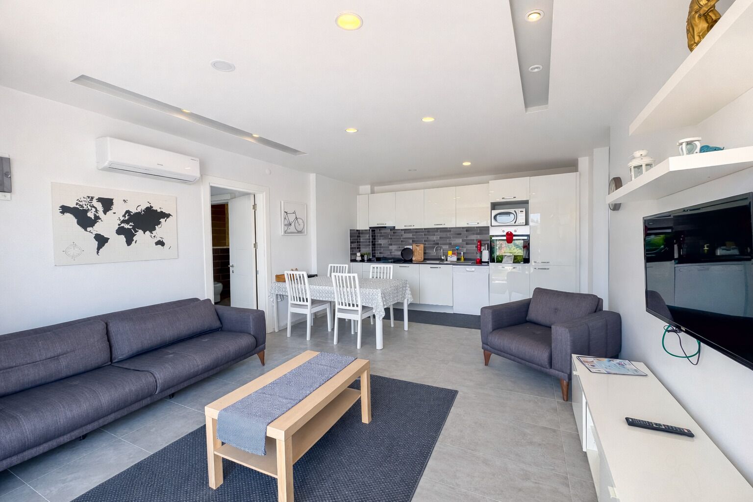 Un apartamento moderno con dos sofás grises, un banco de madera, una mesa de comedor con sillas blancas, una zona de cocina y un televisor montado en la pared.
