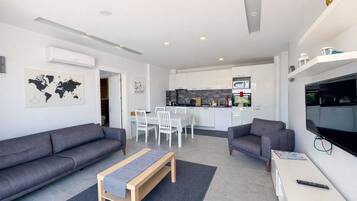 Un apartamento moderno con dos sofás grises, un banco de madera, una mesa de comedor con sillas blancas, una zona de cocina y un televisor montado en la pared.