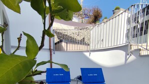 A terrace with two blue lounge chairs labeled "ALISEA HOME" and a small white table, surrounded by a white wall and a stone wall in the background.