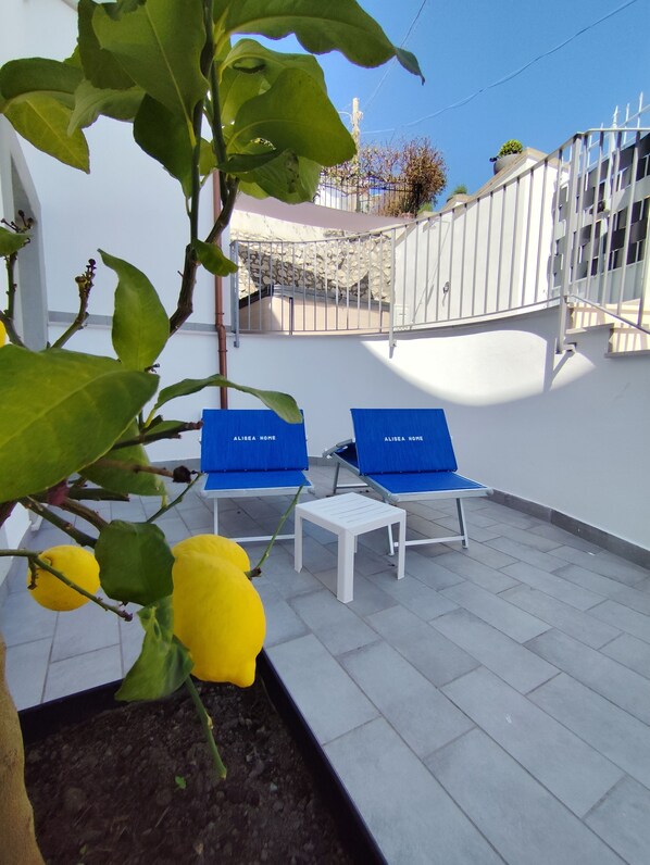 Une terrasse avec deux fauteuils bleus, portant l’inscription « ALISEA HOME », et une petite table blanche, le tout entouré d’un mur blanc et, au fond, d’un mur en pierre.