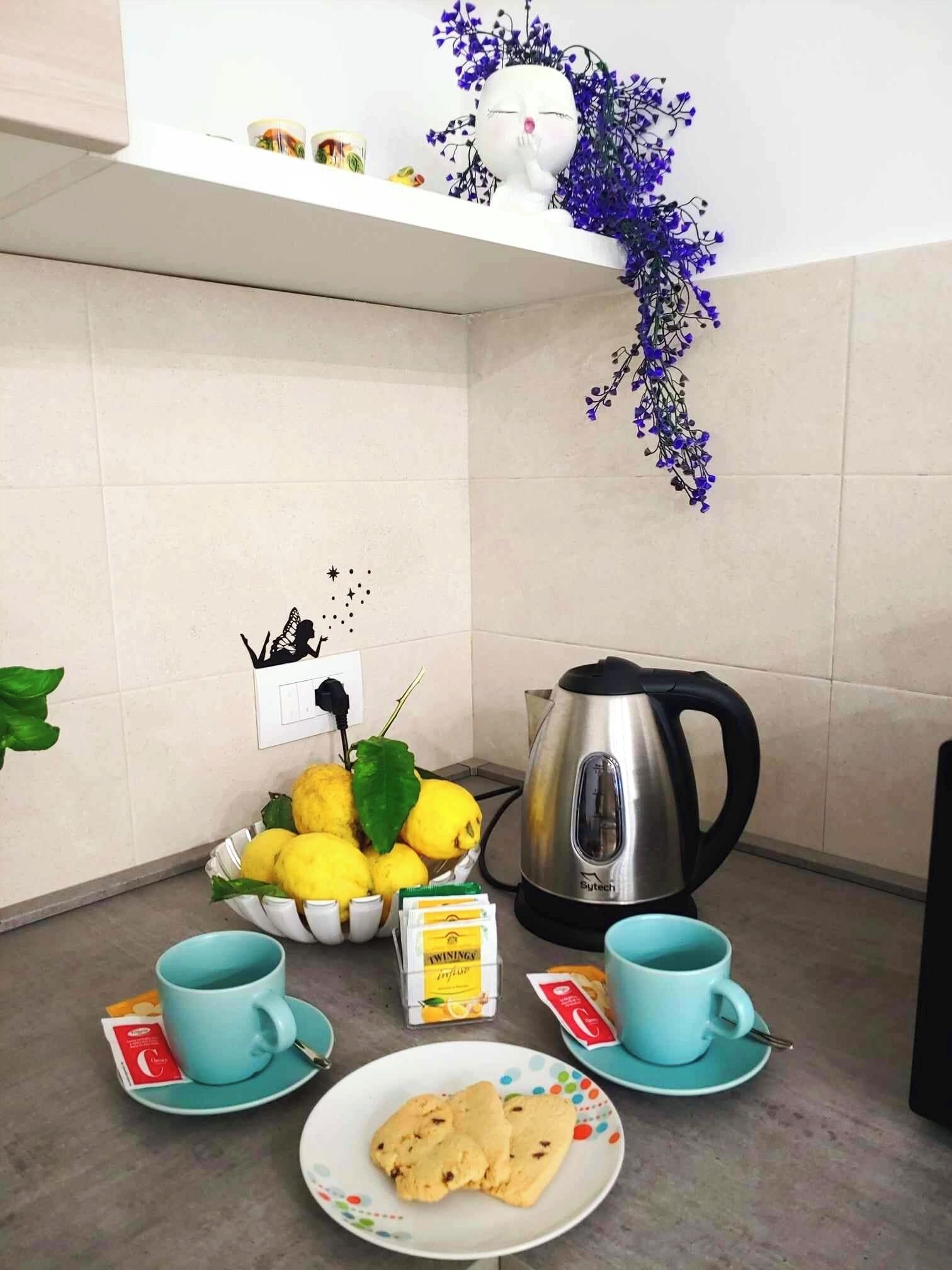 A kitchen with a kettle, teacups, a plate of cookies, lemons, and a decorative plant.