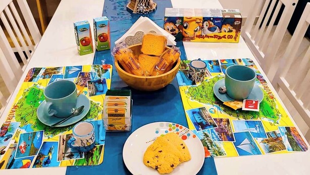 Una mesa de comedor con mantel azul, un bol de galletas, un plato de galletas, una taza, un platillo y una tetera. También hay algunas cajas y un pequeño objeto decorativo sobre la mesa.