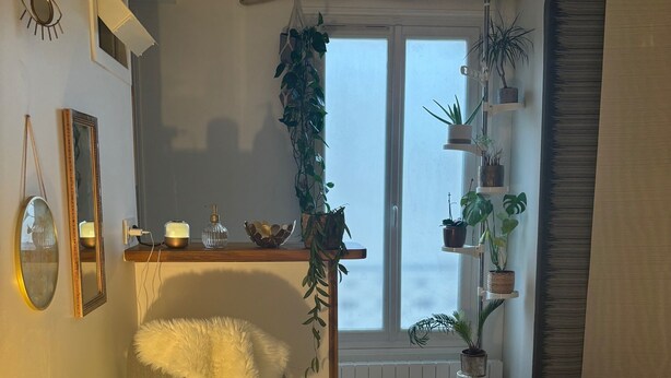 A cozy living room with a white sofa, a dining table with a lamp, and a large window with plants. - Studio Cosy et Tout Confort à Proximité Immédiate du Canal ! Cœur de Paris (Paris)