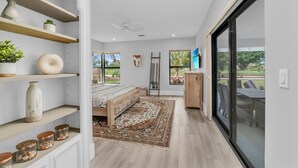 A modern bedroom with a bed, shelves with decorative items, a large window, and a view of the outdoors. - Luxury Golf-View Oasis with Heated Pool & Quiet Marco Island Location (Marco Island)