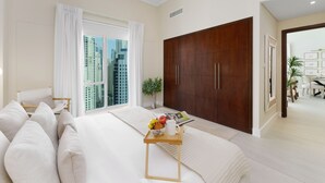 A modern bedroom with a large bed, a wooden wardrobe, a bedside table with a tray, and a view of the city through a large window. - Maison Privee - Modern Beach Living in the Heart of Dubai Marina (Dubai)