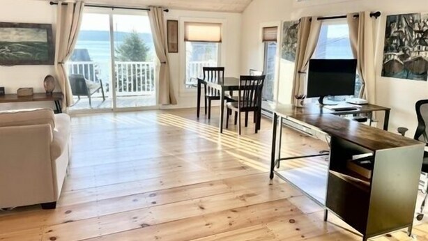 A spacious room with a wooden floor, a dining area with chairs and a table, a sofa, a TV, and a balcony with a view of the sea. - 2 bedroom w/ expansive water views (Phippsburg)