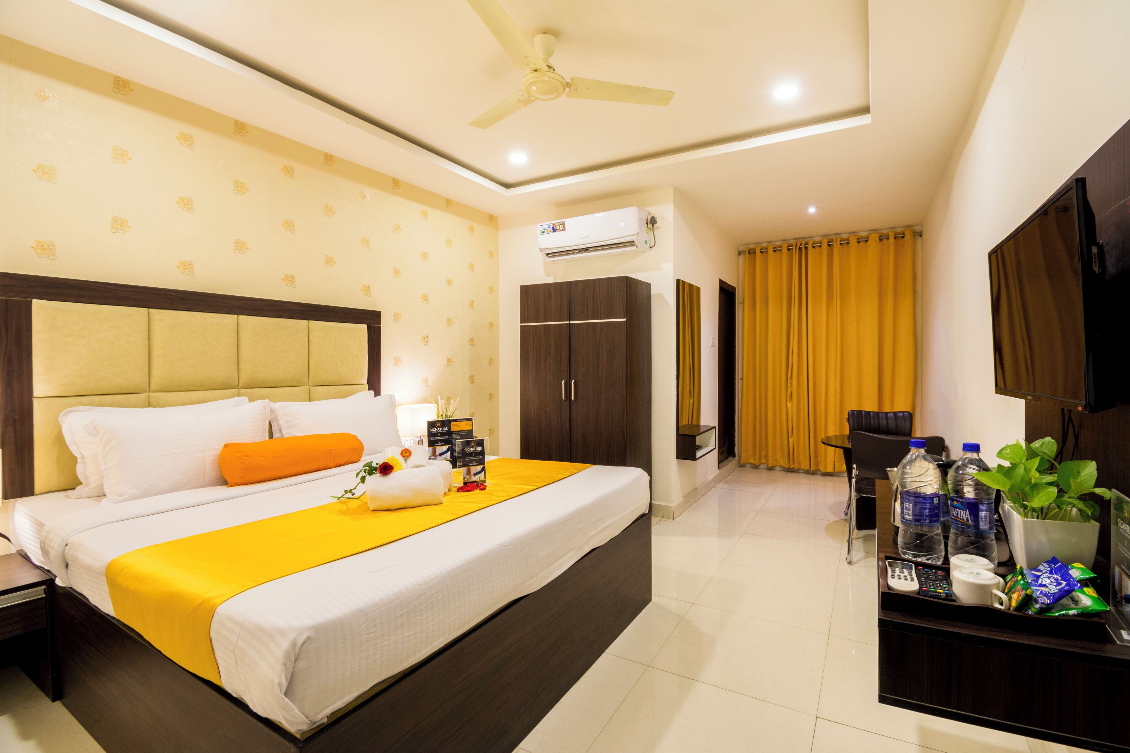A hotel room with a bed, a desk with a TV, a ceiling fan, an air conditioner, and a window with curtains.