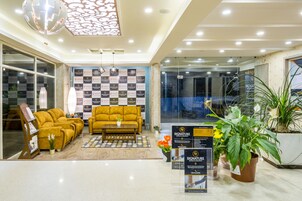 Un hall de hotel con suelo a cuadros, un sofá amarillo, una mesa de centro y plantas en macetas.