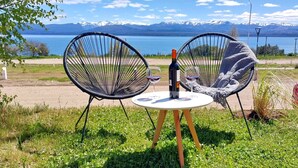 Dos sillas de mimbre negro con una mesa blanca y una botella de vino entre ellas, con vista a un lago y montañas al fondo.