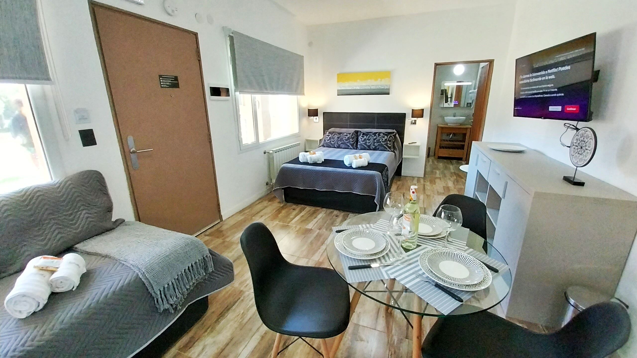 Habitación de hotel con cama, sofá, mesa de comedor para dos personas y televisión montada en la pared.