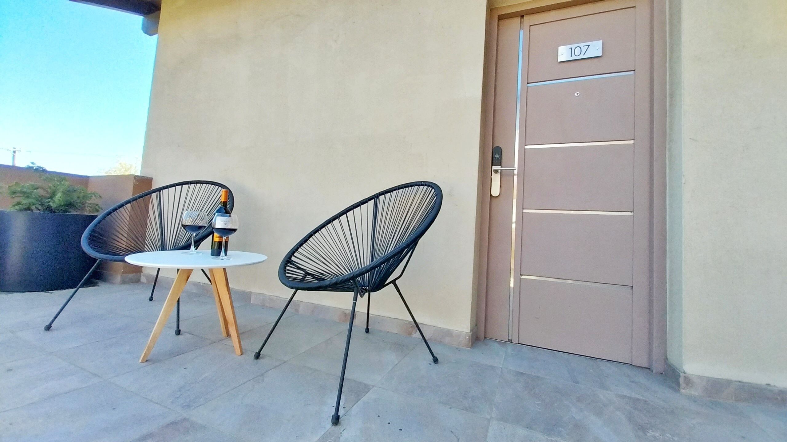 Un área de estar al aire libre con dos sillas y una mesita, una puerta con el número 107, y una planta en maceta al fondo.