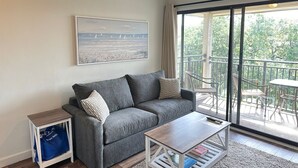A living room with a gray sofa, a coffee table, a painting of a beach, and a balcony with outdoor furniture. - Ocean View, Top Floor Condo in Hilton Head (Hilton Head Island)