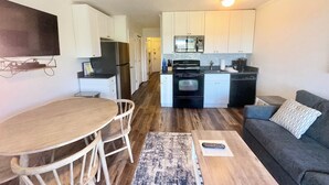 A compact kitchen with white cabinets, a black refrigerator, and a microwave. There's a dining area with a wooden table and chairs, and a gray couch with a patterned pillow. - Ocean View, Top Floor Condo in Hilton Head (Hilton Head Island)