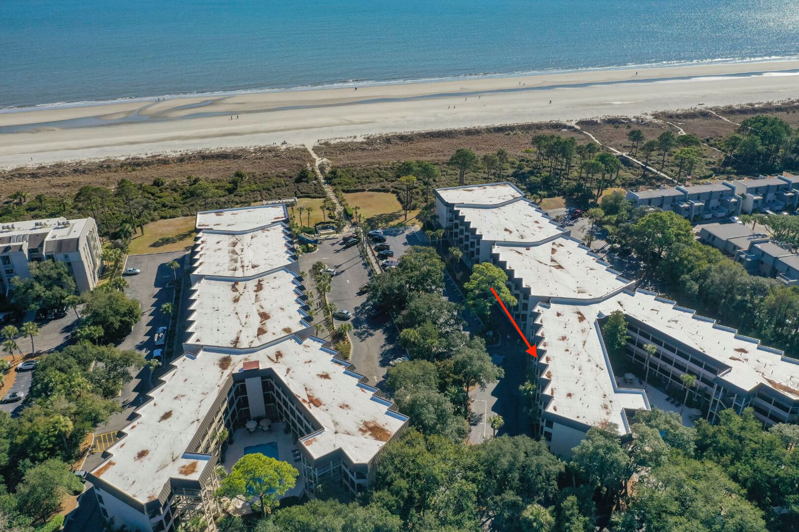 Vue aérienne d’un hôtel situé sur la côte, composé de plusieurs bâtiments, avec un parking et une plage en arrière-plan.