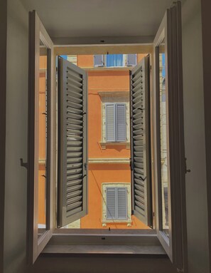 Blick durch ein offenes Fenster mit Fensterläden, das ein Gebäude mit orangenen Wänden und einem Balkon zeigt.