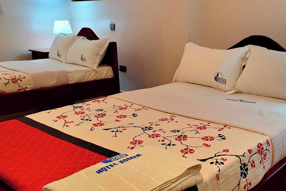 A hotel room with two beds, each with a floral bedspread and pillows featuring the hotel's logo. There is a bedside table with a lamp and a red throw blanket.