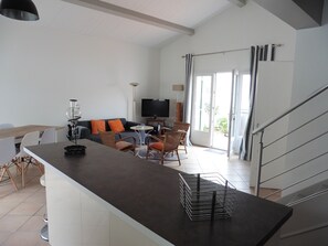 A spacious living area with a kitchen counter, dining table, and seating. A flat-screen TV is mounted on the wall, and a staircase leads to an upper level.
