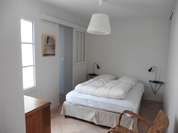 A bedroom with a bed, two bedside lamps, a wooden nightstand, a wicker chair, and a hanging light fixture.