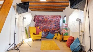 Een gezellige kamer met een metselwerkmuur, een gele fauteuil, kleurrijke kussens en fotografische apparatuur.