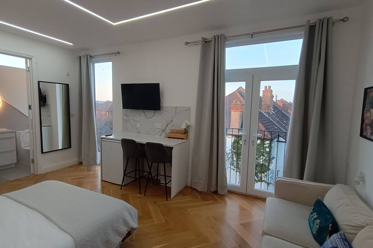 A modern hotel room with a large bed, a desk with two chairs, a TV, and a balcony with a view of buildings.