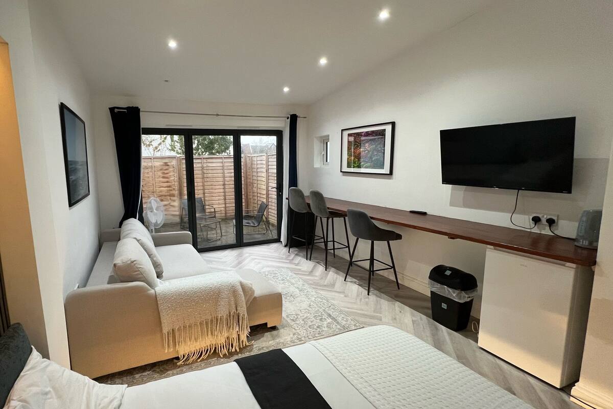 A modern hotel room with a large bed, a sofa, a desk with two chairs, a TV, and a sliding glass door to the outside.