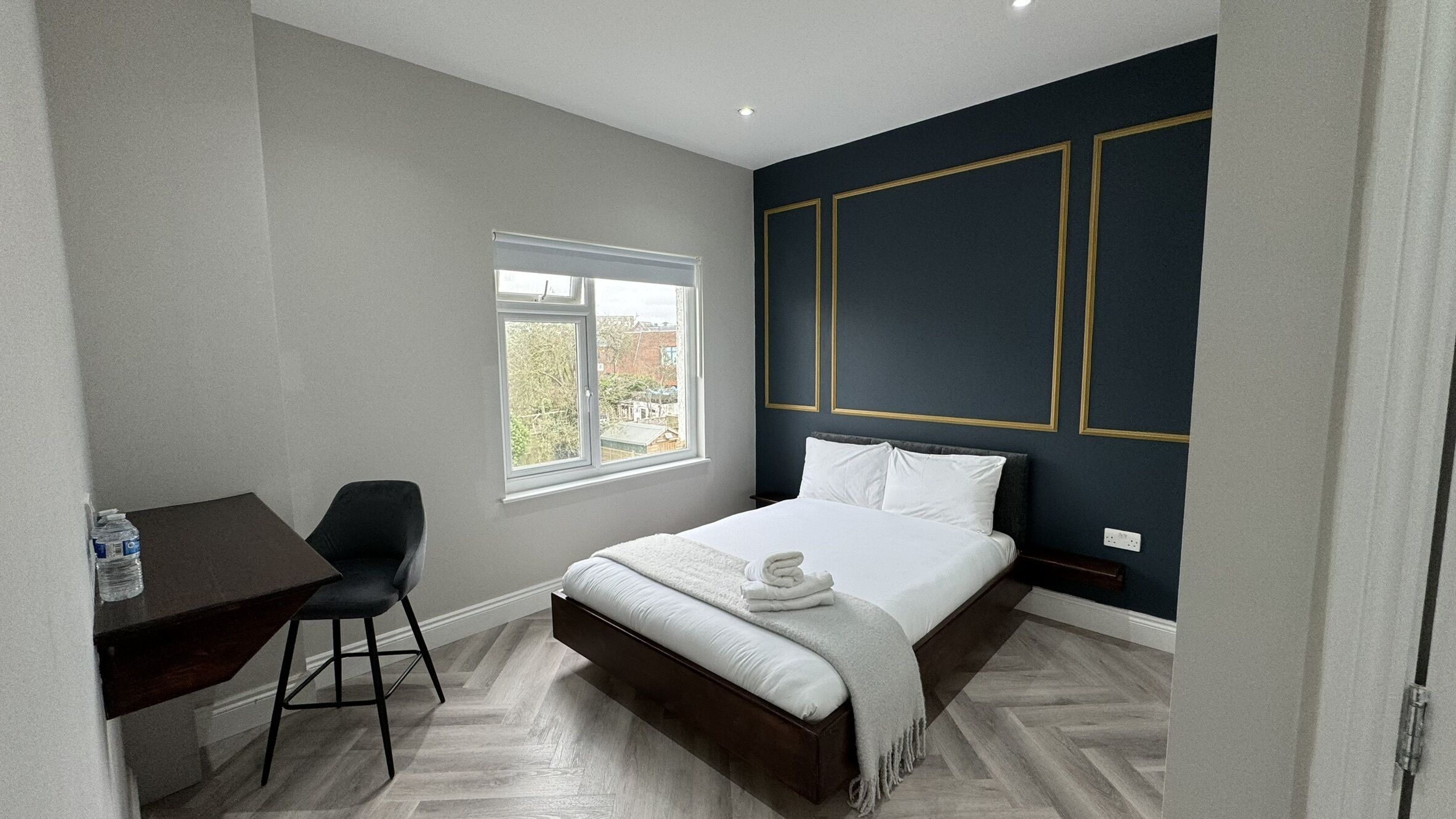 A bedroom with a bed, a desk, and a chair. There is a window with a view of the outside.