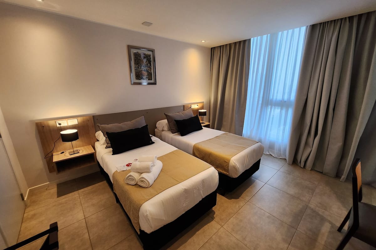 Hotellihuone, jossa on kaksi sänkyä, yöpöytä lampun kanssa, seinällä oleva kehystetty kuva ja suuri ikkuna, jossa on verhot.