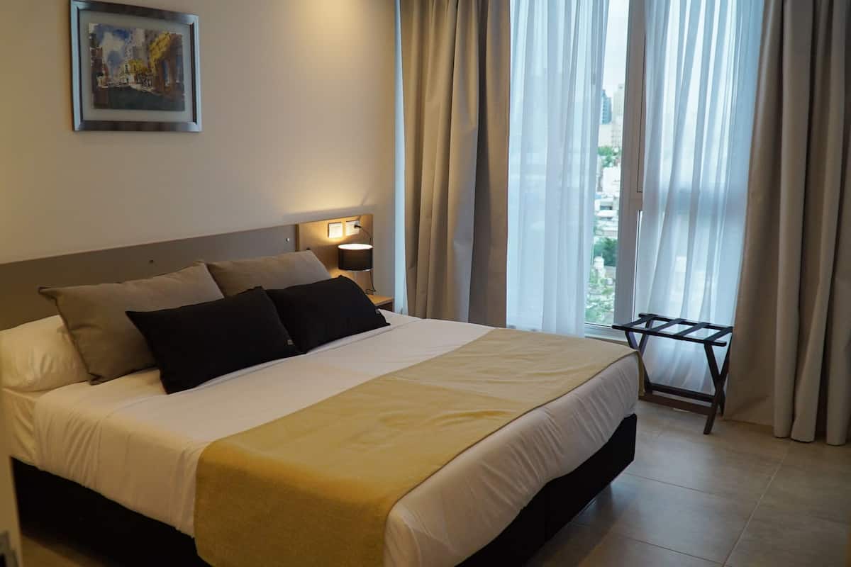 Hotellihuone, jossa on suuri sänky, yöpöytä lampulla, ikkuna verhoilla ja näkymä kaupunkiin.