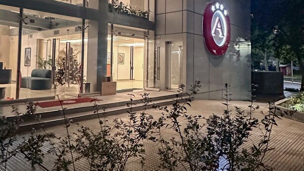 A modern building entrance with a glass facade, a prominent logo on the wall, and a well-lit pathway leading to the entrance. - Amerian Rio Cuarto (Rio Cuarto)