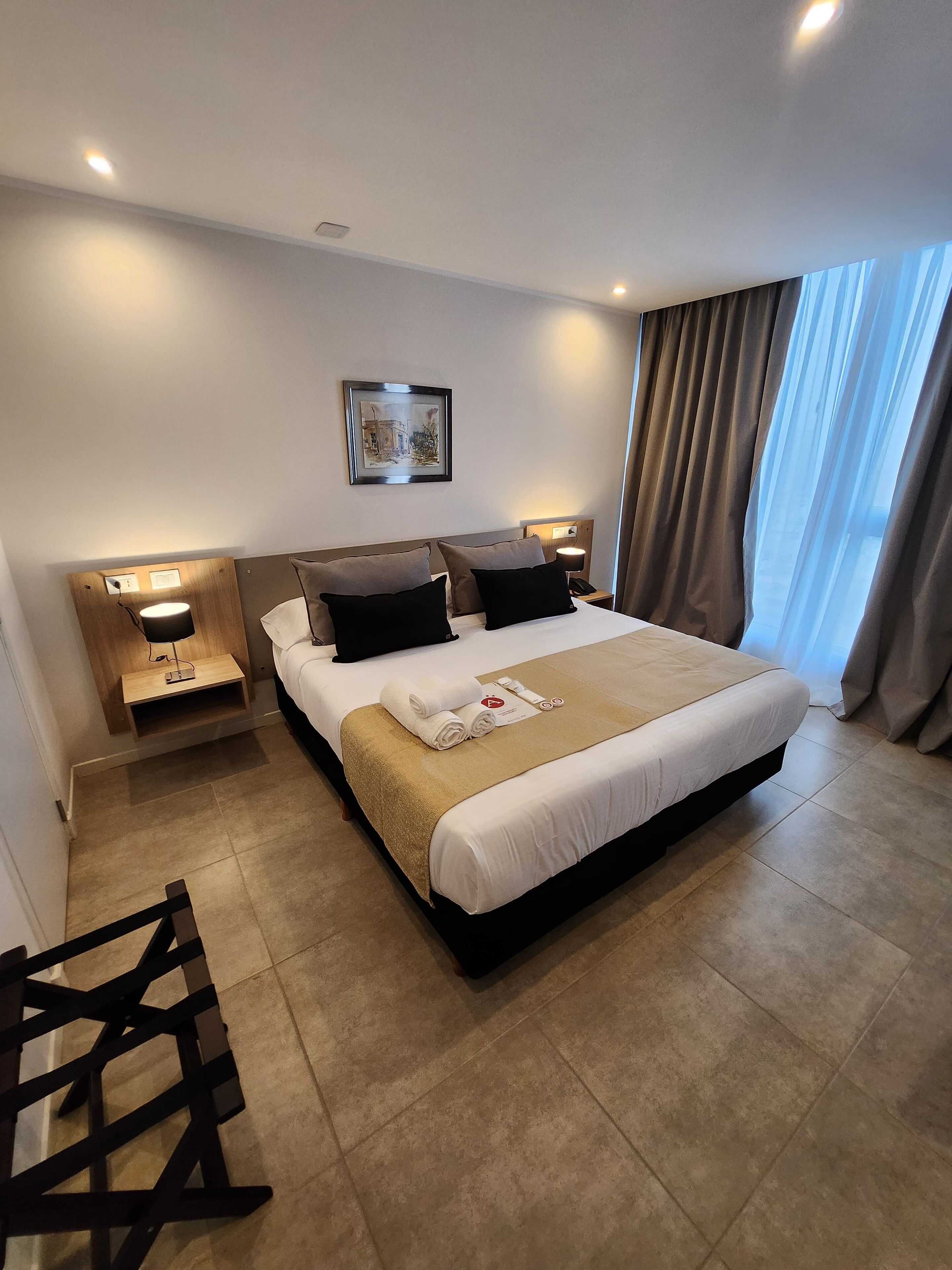 A hotel room with a large bed, a bedside table with a lamp, a framed picture on the wall, and a window with curtains.