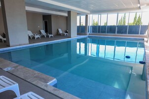 Una piscina interior con agua azul cristalina, suelo de baldosas y sillones blancos.