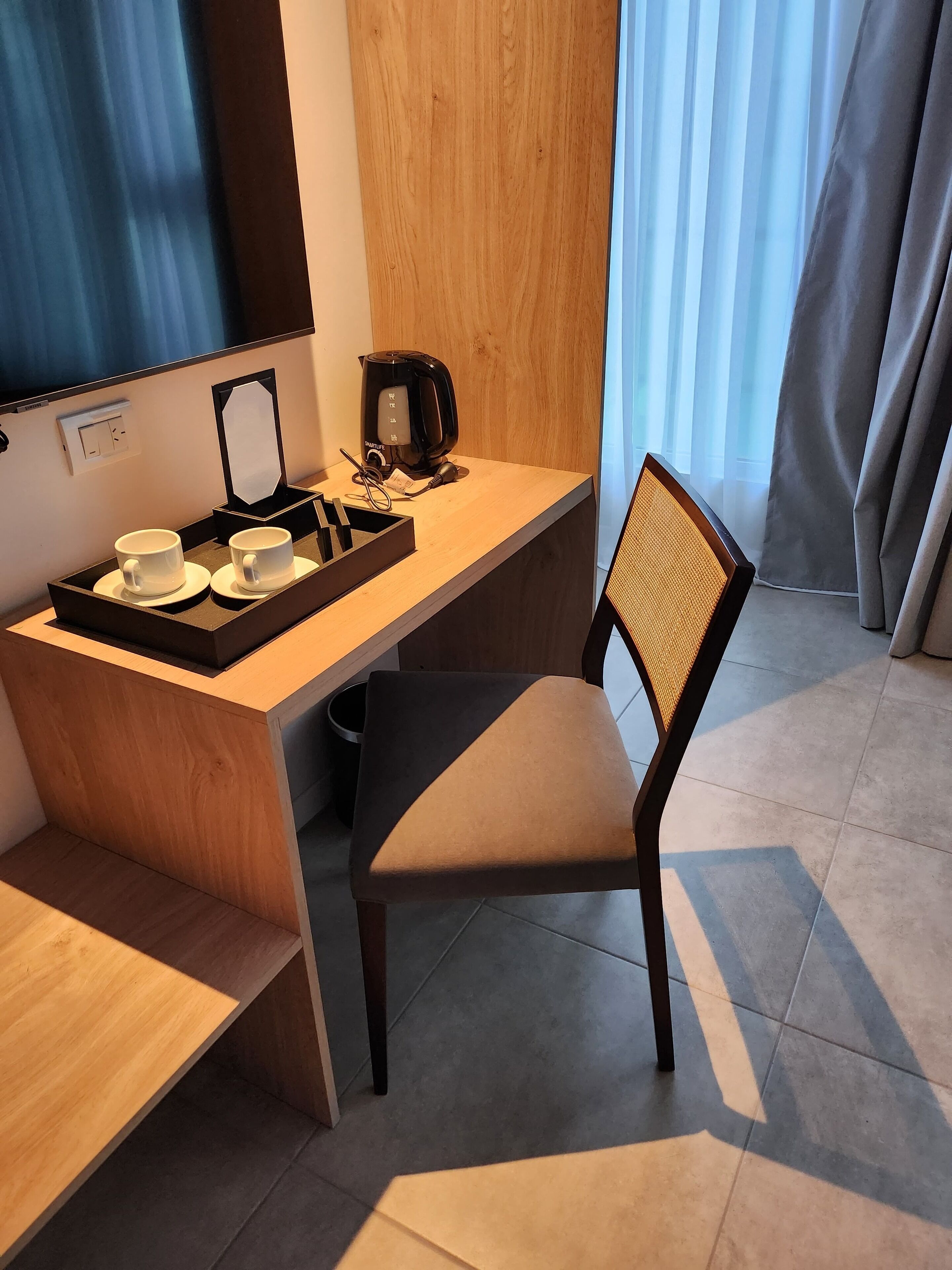 Una habitación de hotel compacta con un escritorio de madera, un hervidor eléctrico negro, dos tazas blancas y una silla con respaldo trenzado.
