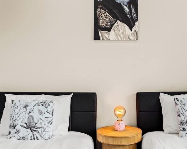 Une chambre avec deux lits, chacun agrémenté d’un coussin à motifs floraux, une table de chevet ronde en bois avec une lampe, et une grande peinture abstraite accrochée au mur.