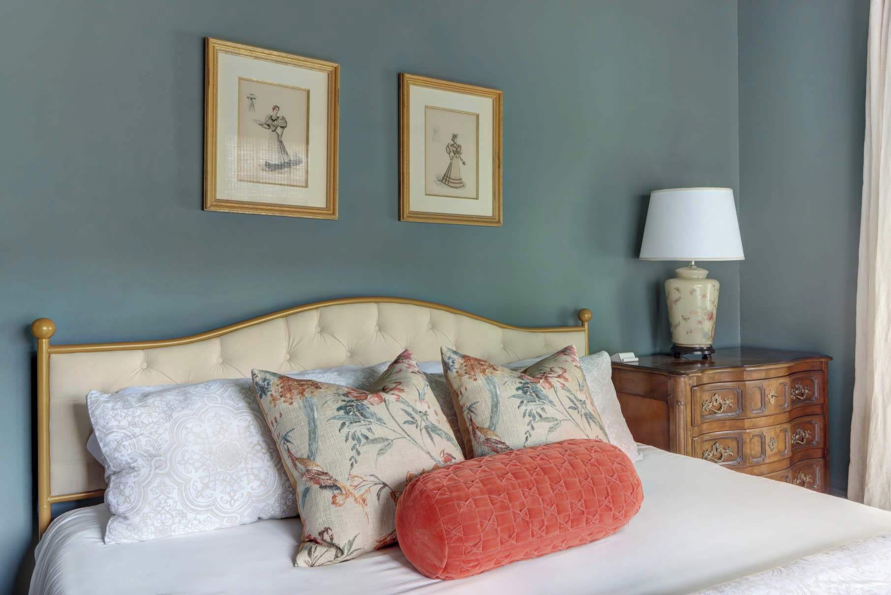 A bed with floral pillows, a wooden nightstand with a lamp, and two framed pictures on the wall.