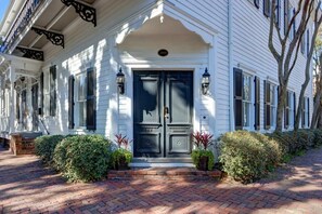 A white building with a black front door, two windows with shutters, and a brick pathway leading to the entrance. - The Gaelic Guesthouse @ Washington Square | 1890s (Savannah)