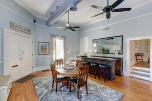 A dining area with a wooden table, chairs, and a ceiling fan. There is a kitchen area with a bar and a large mirror. - The Gaelic Guesthouse @ Washington Square | 1890s (Savannah)