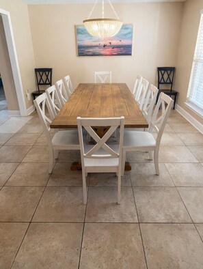 A dining room with a wooden table, white chairs, a chandelier, and a sunset painting on the wall.