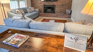 A living room with a brick fireplace, a flat-screen TV, a grey sofa, and a coffee table with a book and a sign reading "wifi".