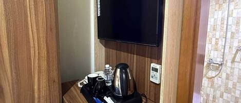 Ein kompaktes Hotelzimmer mit Schreibtisch aus Holz, einem Flachbildfernseher, einer Kaffeemaschine und einem kleinen Tablett mit Tassen und einer Flasche.