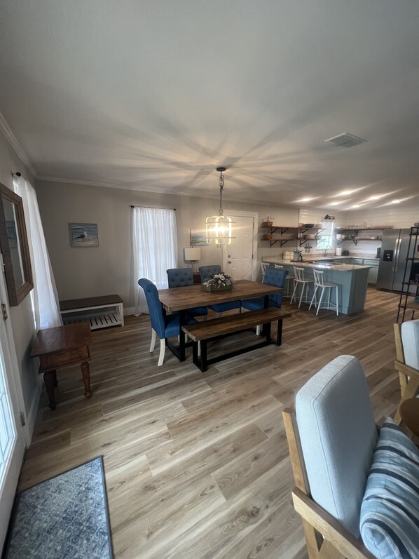 Een ruime woonkamer met een houten eettafel, blauwe stoelen, een bankje en een keuken met een koelkast en spoelbak.