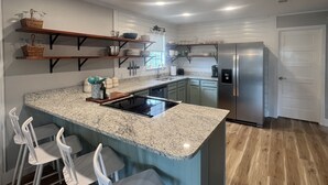 Une cuisine dotée d’un plan de travail en granit, de tabourets de bar blancs, d’un réfrigérateur en acier inoxydable et d’un revêtement de sol en bois.
