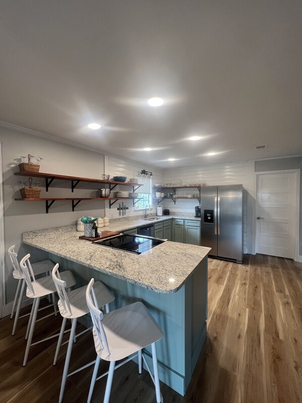 A kitchen with a granite countertop, white bar stools, a stainless steel refrigerator, and wooden flooring.