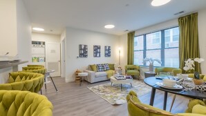 Ein modernes Wohnzimmer mit einer Couch, einem Couchtisch und Sesseln. Es gibt einen Esstisch mit einer Vase und einer Schüssel. Das Zimmer hat ein großes Fenster mit Vorhängen und Blick ins Freie.